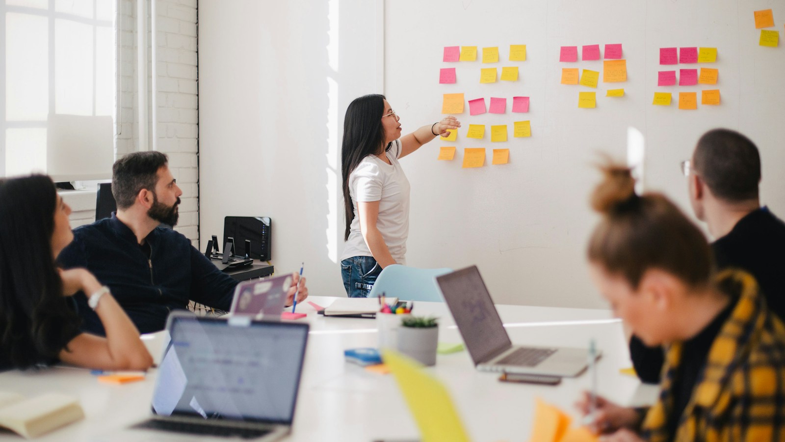 Small business strategy meeting in a sunlit conference room with a laptop presenting a 90-day phased AI adoption roadmap: internal productivity, customer messaging, and automated workflows.